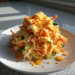 A close-up of a mound of creamy kani salad mixed with shredded carrots and cucumber chunks on a white plate.