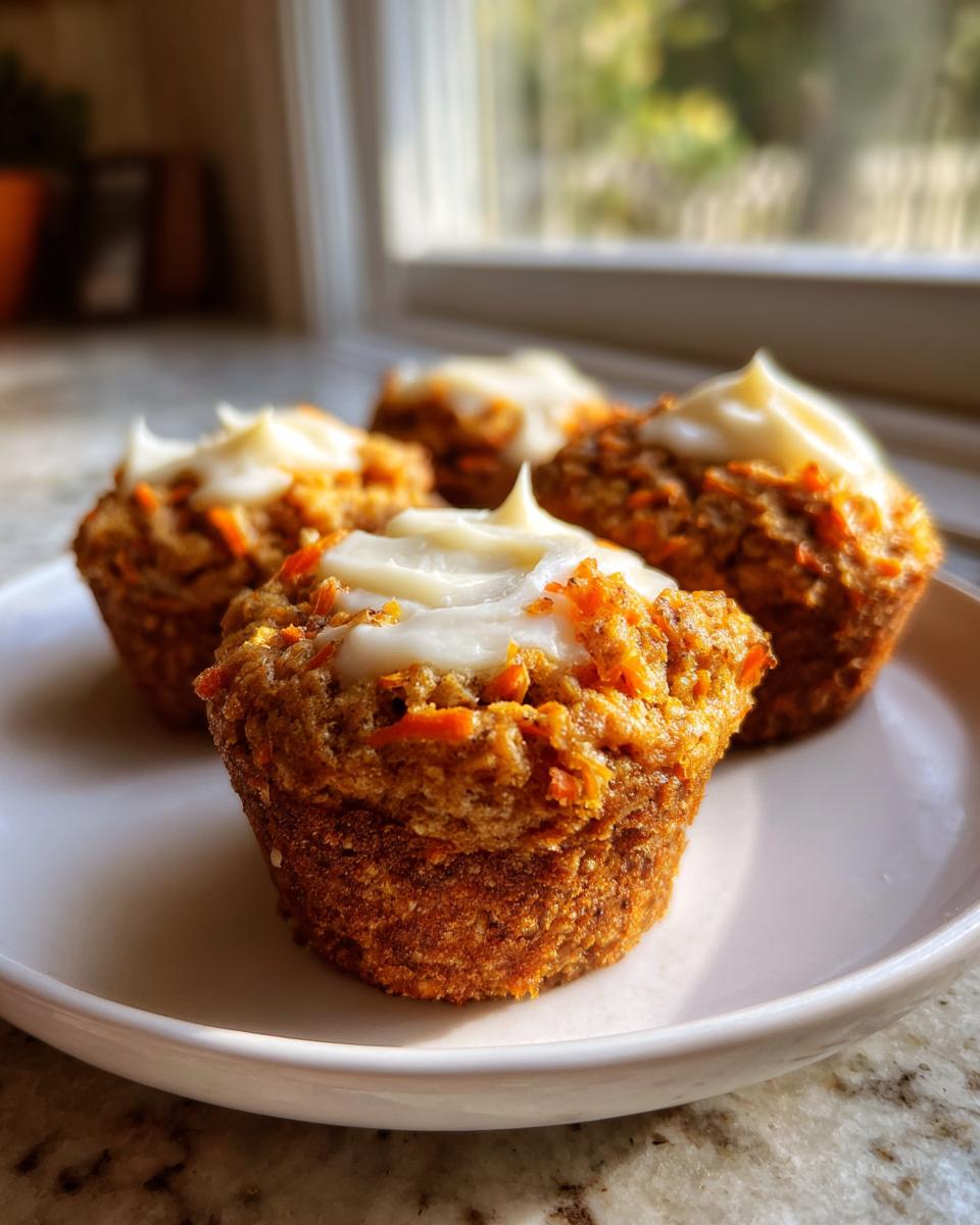 Four moist carrot cake muffins topped with swirls of cream cheese frosting, sitting on a white plate.