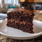 A close-up of a rich, dark slice of moist chocolate cake with thick chocolate frosting on a white plate.