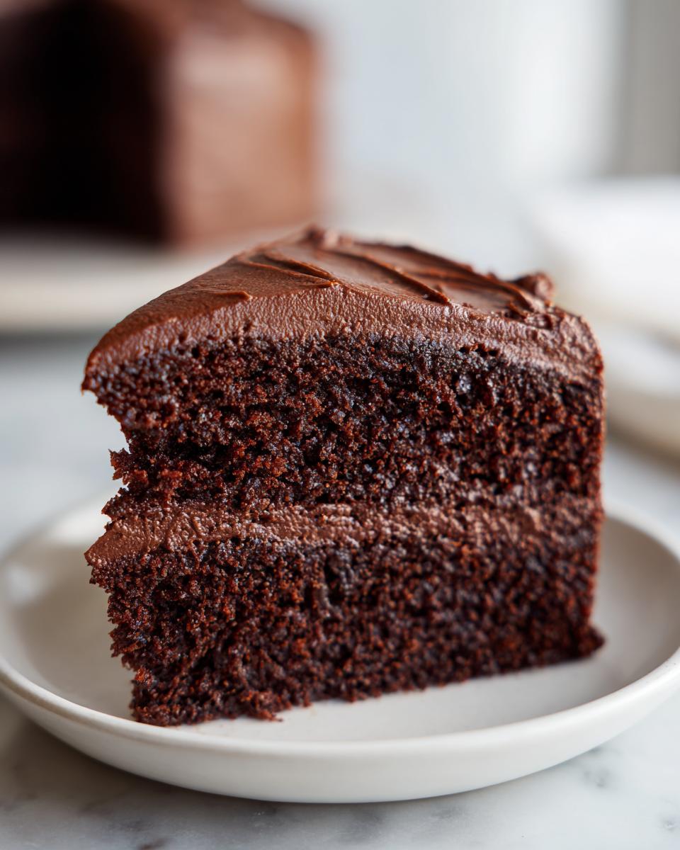Close-up of a moist slice of chocolate cake with thick, dark chocolate frosting on a white plate.