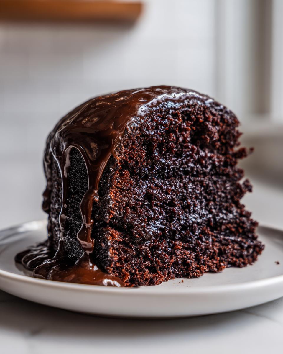 A close-up of a slice of incredibly moist chocolate cake topped with rich, flowing chocolate ganache.