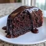 A close-up of a slice of moist chocolate cake topped with thick, glossy chocolate glaze dripping down the side.