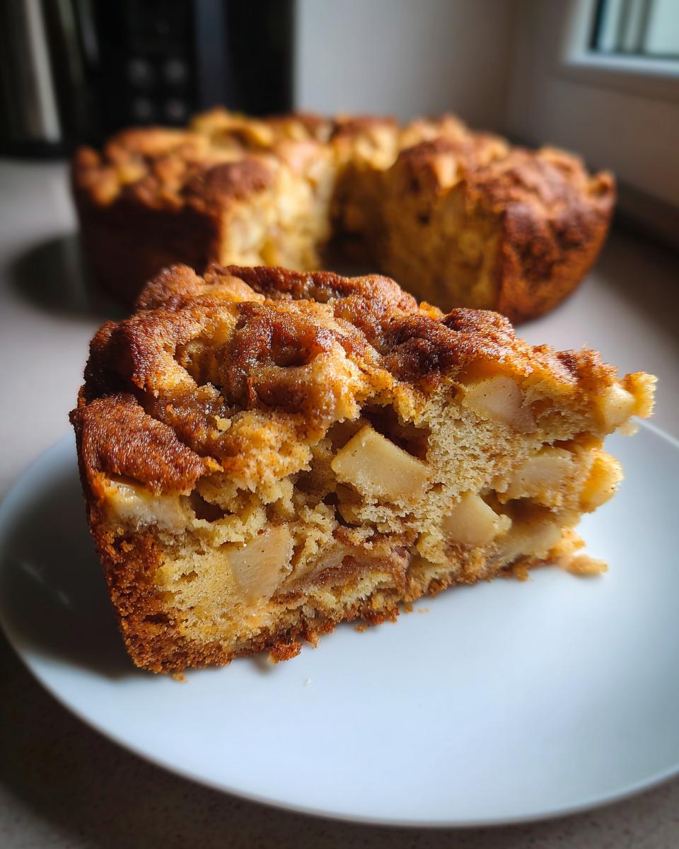 A close-up slice of moist jewish apple cake showing chunks of apple baked into the crumb, with the rest of the cake blurred in the background.