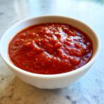 A close-up shot of thick, vibrant red, no-cook pizza sauce seasoned with herbs and pepper, served in a small white bowl.