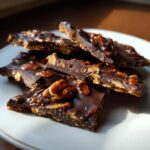 Close-up of broken pieces of dark chocolate pecan pie bark stacked on a white plate, glistening under sunlight.