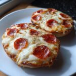 Two perfectly baked pepperoni pizza bagels topped with melted mozzarella cheese and cracked black pepper on a white plate.