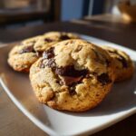 Close-up of three thick, golden brown chocolate chip cookies with melted dark chocolate chunks on a white plate.