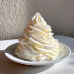 A tall, perfectly piped swirl of white cream cheese frosting sitting on a small white saucer, highlighted by bright sunlight.