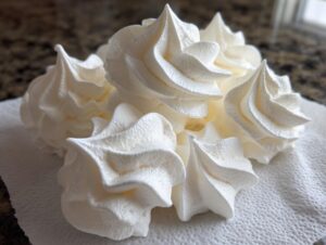 Close-up of several freshly piped, bright white meringue cookies resting on a paper towel.