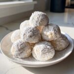 A mound of freshly baked russian tea cakes generously dusted with powdered sugar, stacked on a white plate.