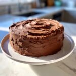 A round layer cake generously covered in thick, swirled chocolate frosting, sitting on a white plate.