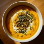 Overhead view of a bowl of vibrant orange roasted butternut squash soup, swirled with cream and topped with pumpkin seeds.