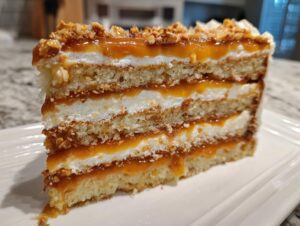 A tall slice of caramel crunch cake showing layers of sponge, white cream, and rich caramel filling topped with nuts.
