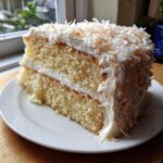 A moist slice of two-layer coconut cake recipe with white frosting and toasted coconut flakes on top.