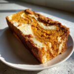 A thick slice of moist pumpkin cream cheese bread showing orange pumpkin cake swirled with white cream cheese filling and a brown streusel top.