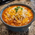 A close-up of a bowl of rich, orange slow cooker buffalo chicken chili topped with shredded cheddar and Monterey Jack cheese and green onions.
