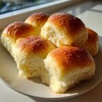 Five fluffy, pull-apart dinner rolls with a golden, buttery glaze stacked on a light plate.