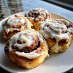 Four freshly baked sourdough discard cinnamon rolls generously topped with white cream cheese icing on a white plate.