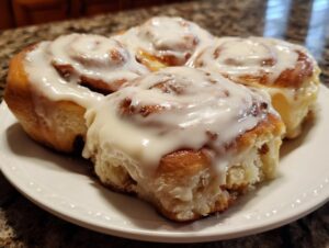 Close-up of four freshly baked sourdough discard cinnamon rolls generously topped with white icing on a white plate.