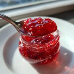 A spoonful of glistening, bright red raspberry jam being lifted from a small glass jar.