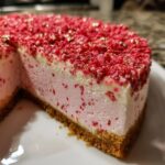 Close-up of a slice of strawberry crunch cheesecake showing the graham cracker crust, pink filling, and red crunch topping.