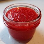 A close-up view of vibrant red strawberry freezer jam filling a small glass jar, showing its textured surface.