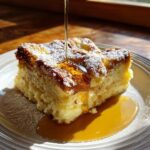 A close-up of a square slice of baked french toast topped with powdered sugar and rich syrup being poured over it.