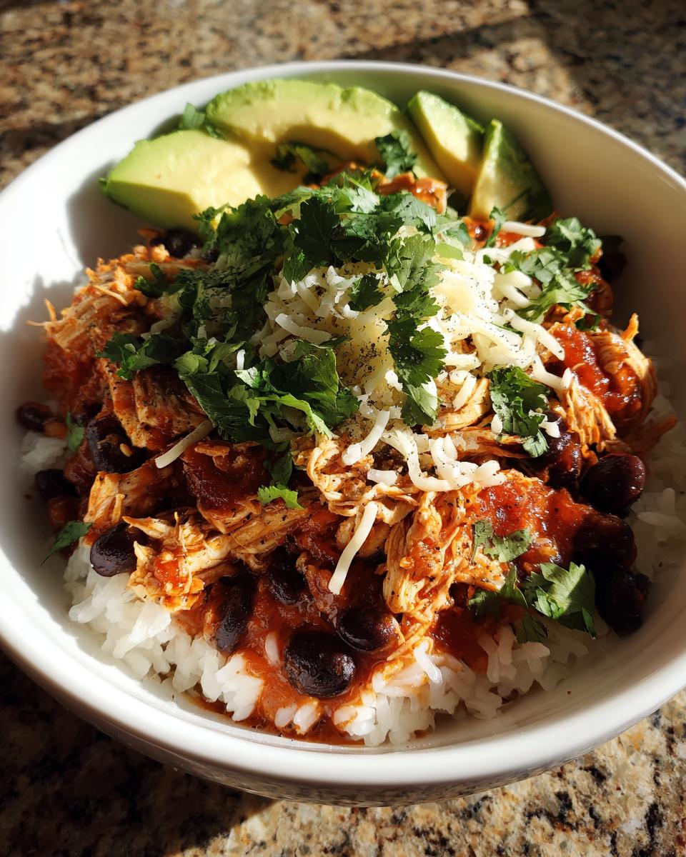 A vibrant chicken burrito bowl featuring shredded chicken, black beans, rice, topped with cheese, cilantro, and avocado slices.