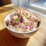 A close-up photo of creamy salmon salad mixed with red onion and celery in a white bowl.