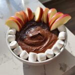 A white bowl filled with rich, dark chocolate dip, garnished with apple slices and mini marshmallows.