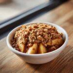 A close-up of a white bowl filled with warm apple crisp recipe, featuring tender apples and a thick, crumbly topping.