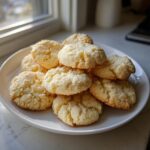 A pile of freshly baked, pale yellow butter cookies with a slightly crinkled texture stacked on a white plate near a window.