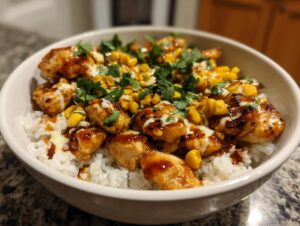 A close-up of a white bowl filled with a delicious chicken rice bowl, topped with glazed chicken pieces, corn, and cilantro.