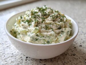 A close-up of creamy dill pickle dip topped with fresh dill and black pepper in a white bowl.