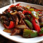 A close-up serving of glossy, saucy pepper steak mixed with bright red and green bell peppers and onions.