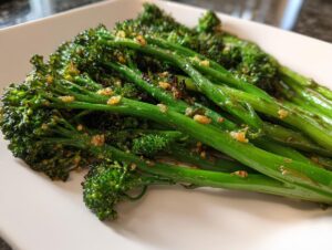 Close-up of bright green roasted broccolini stalks seasoned with crispy garlic bits on a white plate.