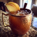 A frosty copper mug filled with an apple cider moscow mule, garnished with apple slices and a cinnamon stick.