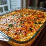 A close-up of freshly baked street corn dip in a clear glass baking dish, topped with melted cheese, cilantro, and chili flakes.