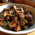 Close-up of a bowl of glossy chicken zucchini stir fry coated in dark sauce and topped with white sesame seeds.