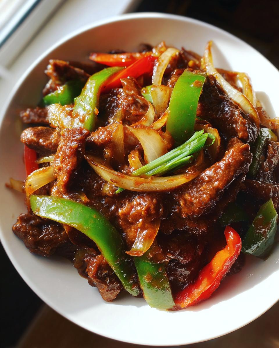 A close-up of a white bowl filled with rich, saucy chinese pepper steak, featuring strips of beef, green and red bell peppers, and onions.