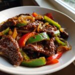 Close-up of glossy, saucy chinese pepper steak with red and green bell peppers and onions in a white bowl.