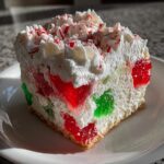 A festive slice of Christmas poke cake featuring red and green gelatin cubes, white whipped topping, and crushed peppermint candies.