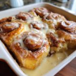 Close-up of gooey cinnamon rolls with heavy cream glaze soaking in a white baking dish.