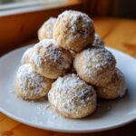 A stack of freshly baked cinnamon sugar snowball cookies heavily dusted with powdered sugar on a white plate.