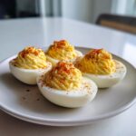Four halves of classic deviled eggs with creamy filling piped high and sprinkled with paprika on a light plate.