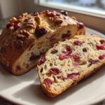 A loaf of freshly baked cranberry walnut bread, partially sliced, showing dried cranberries and walnuts inside.