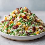 A mound of creamy pea salad mixed with cheddar cheese cubes and topped with crispy bacon pieces.