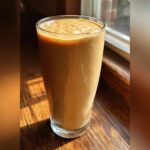 A tall glass filled with a thick, creamy banana smoothie, sitting on a wooden surface near a window.
