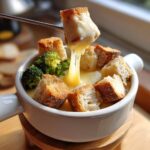 A piece of bread being dipped into a pot of creamy cheese fondue recipe, showing long cheese pulls.