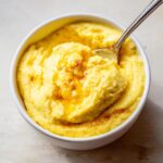 Close-up overhead shot of creamy, yellow cheese grits in a white bowl with a spoonful being lifted and melted butter drizzled on top.